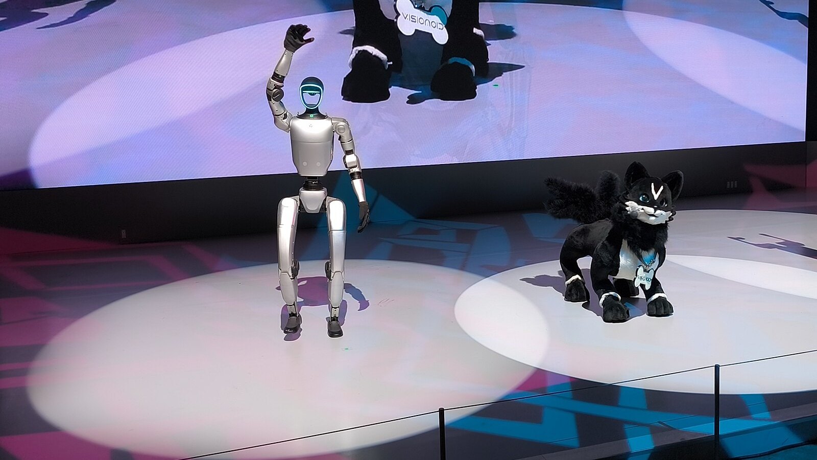 Silberner humanoider Roboter winkt auf einer beleuchteten Messebühne, daneben ein schwarzer Plüsch-Roboterhund