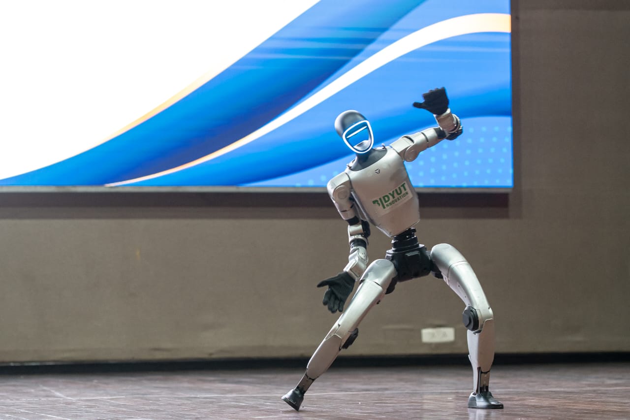 Silberner zweibeiniger humanoider Roboter in Ausfallschritt-Pose auf einer Bühne vor einer großen Leinwand