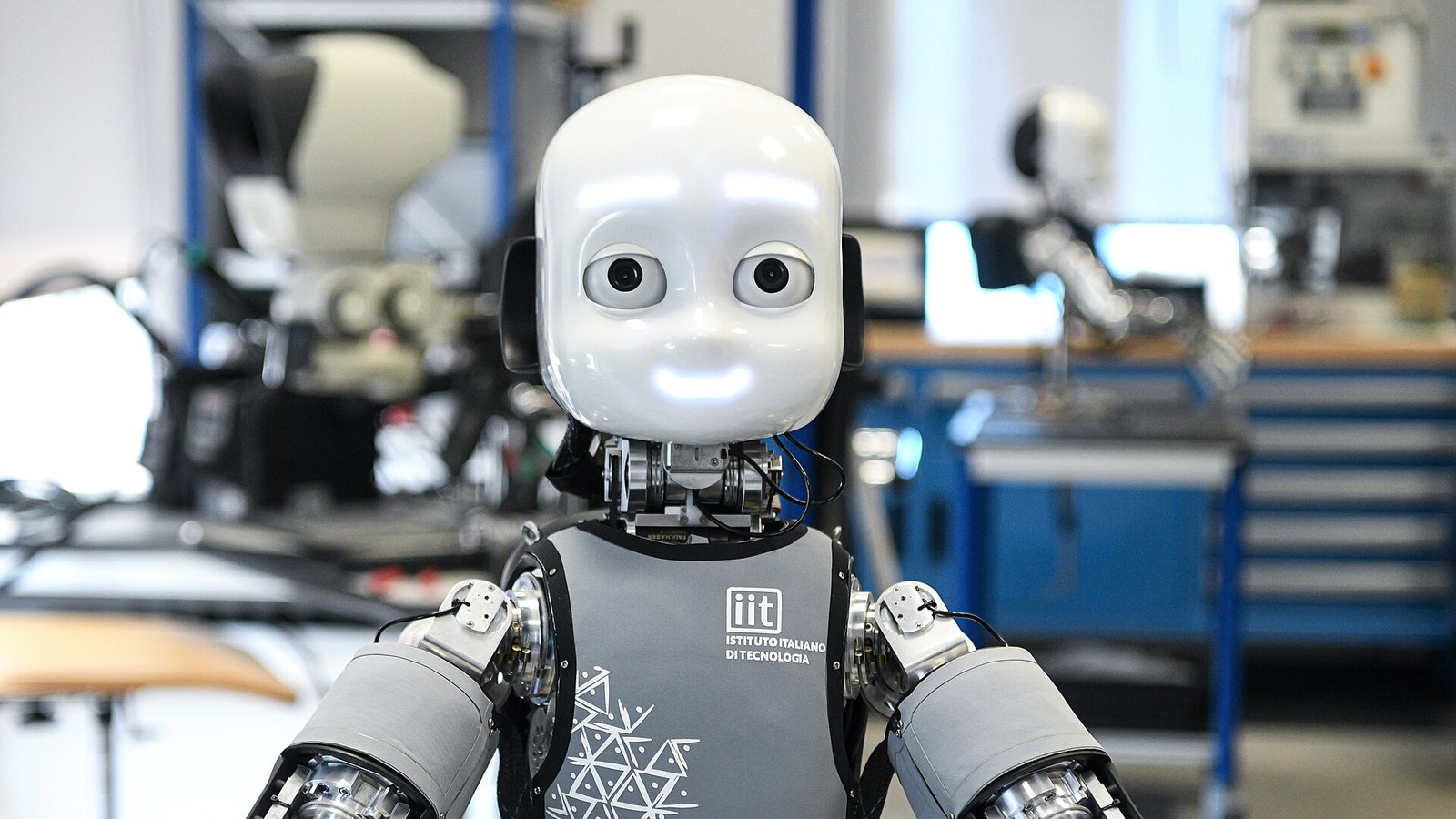 Humanoider Forschungsroboter iCub mit weißem Kopf und großen Augen in einem Robotiklabor, im Hintergrund Werkbänke und weitere Roboterteile